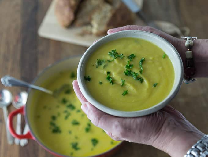 two hands holding a bowl of cauliflower soup with parsley and a dutch oven of soup below