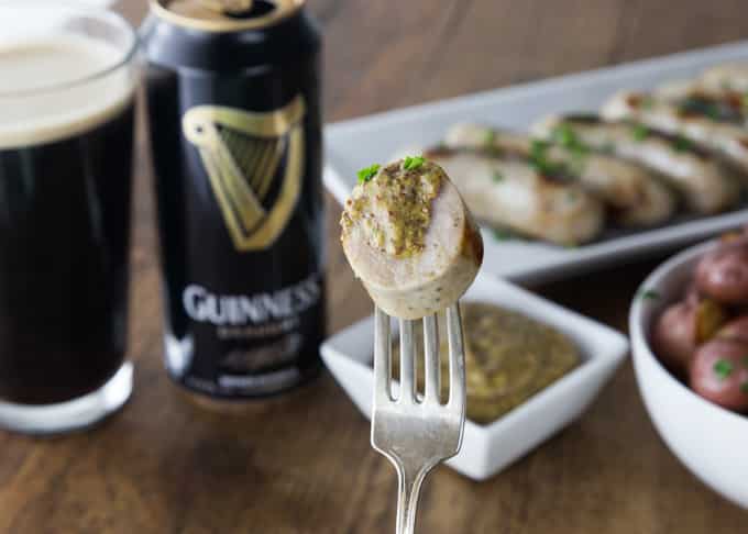 a fork with guinness mustard with a bowl of mustard, sausages, and guinness in the back