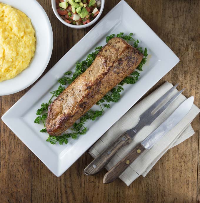 a roasted pork loin on a plate next to a fork and a knife with parsley and a bowl of grits and pico de gallo above 