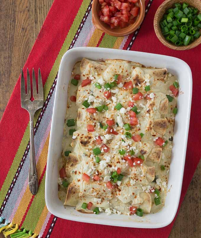 a tray of chicken enchiladas in white sauce topped with jalepenos and tomatoes on a red placemat with a fork on the left
