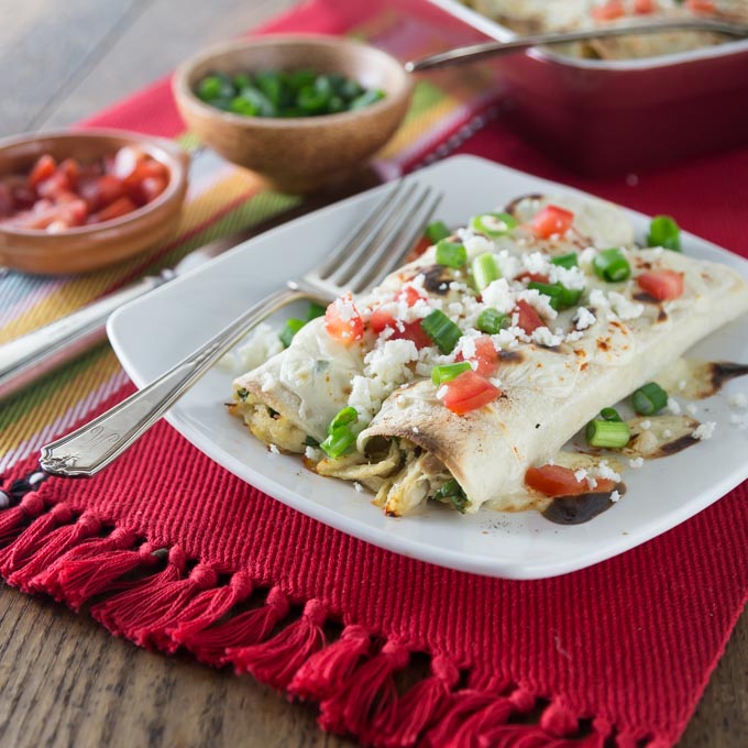 A photo of two chicken enchiladas on a plate with tomato and green onions.