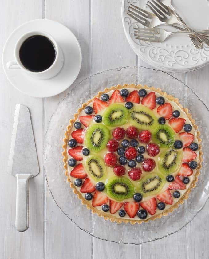 A large fruit topped dessert on a glass plate with forks, a cake knife and coffee