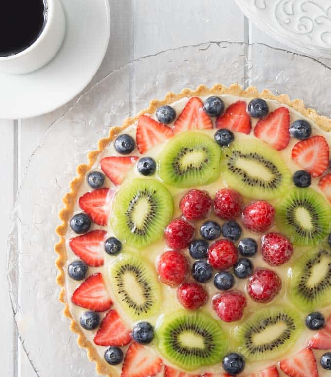 a fruit topped tart on a glass plate with coffee on the left
