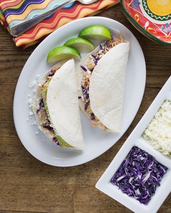 two large chicken tinga tacos on a plate with lime wedges and a bowl of cabbage and cheese on the right