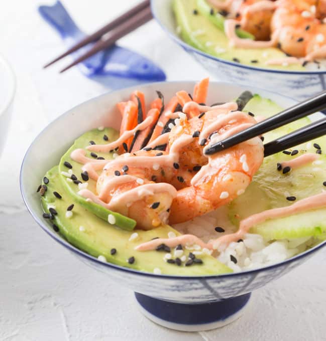 a bowl of rice with sliced avocado, shrimp, and spicy sauce with chopsticks grabbing the shrimp