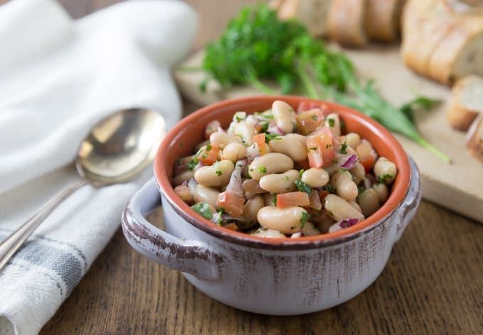 a ceramic dish of cannellini bean salad with sliced bread and herbs in the back