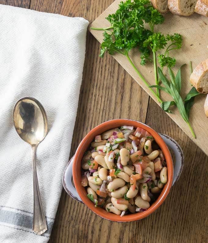 bean salad in a ceramic bowl and a wooden cutting board with herbs and bread on the right