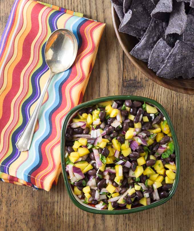a bowl of mango black bean salsa and a bowl of chips with a colored napkin and spoon on the left