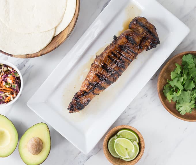 a grilled pork tenderloin on a white plate with taco ingredients around it
