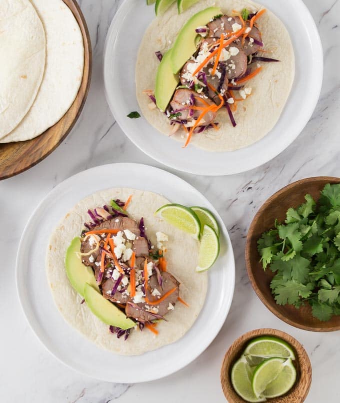 two prepared pork tacos on white plates with taco shells, coriander, and limes around 