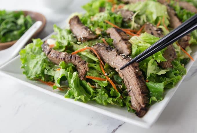 a salad on a square plate with sliced beef and chopsticks 