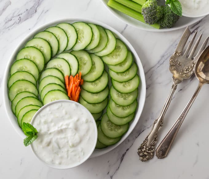 a plate of cucumbers with a bowl of tzatziki with a fork and spoon on the right