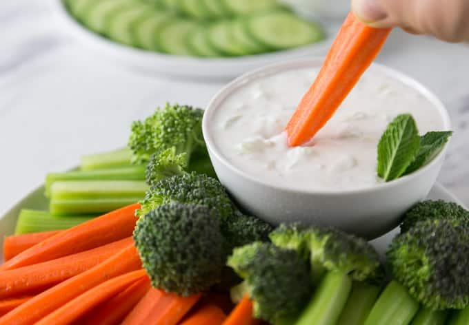 a plate of chopped vegetables with a bowl of tzatziki and a carrot dipping in