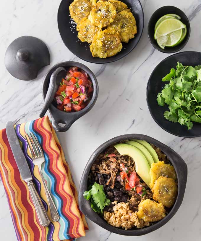 a pork carnitas bowl with fried plantains, rice, beans, and avocado surrounded by plates of cilantro, plantains, salsa and a napkin with a fork and knife