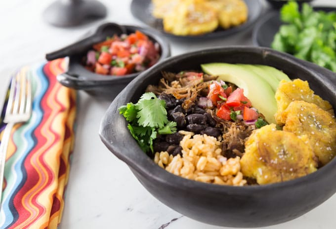 a pork carnita bowl with fried plantain, avocado, salsa, beans, and cilantro with toppings dishes in the back