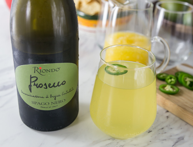 a bottle of prosecco next to a mango cocktail in a glass with a slice of jalepeno