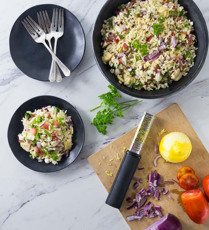 a black bowl of mediterranean orzo pasta salad with a served plate, a plate of forks, and a cutting board with ingredients