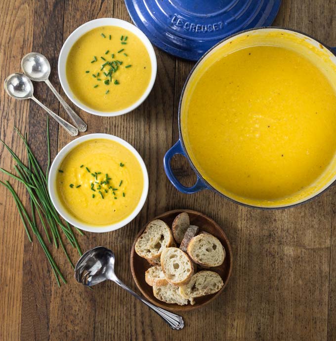 a dutch oven and two bowls of squash soup with chives, spoons, and a bowl of sliced baguette