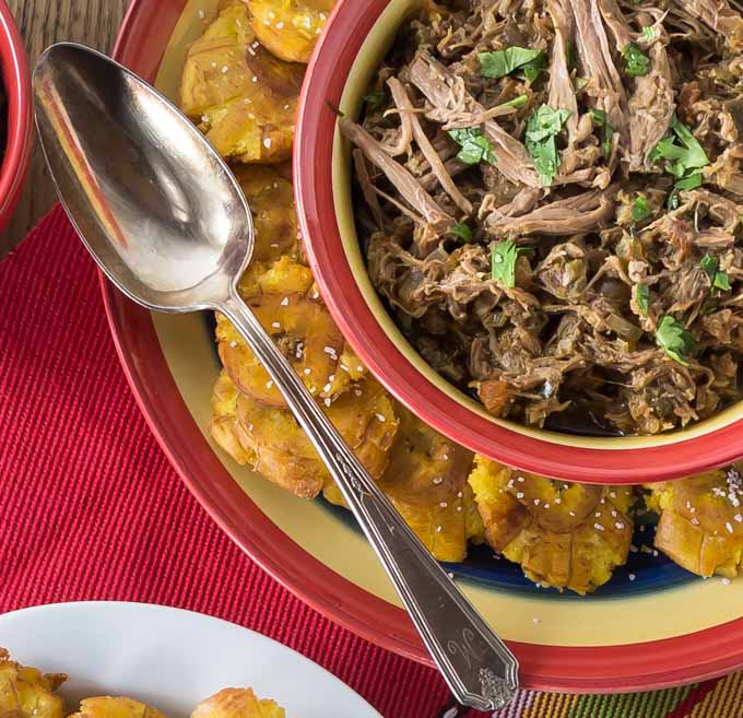 a bowl of ropa vieja with a plate of fried plantains and a spoon