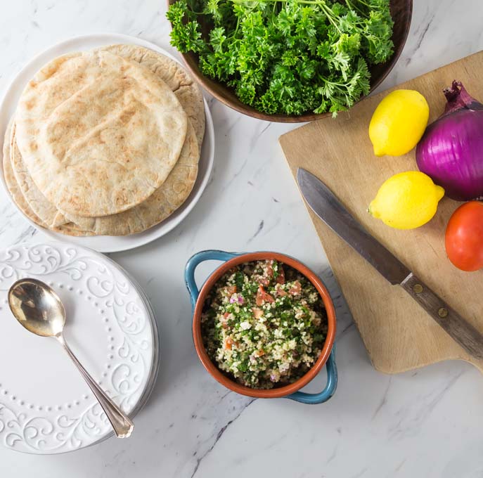 a bowl of tabbouleh, a cutting board with lemons, a bowl of parsley, and a plate of pita on a marble countertop
