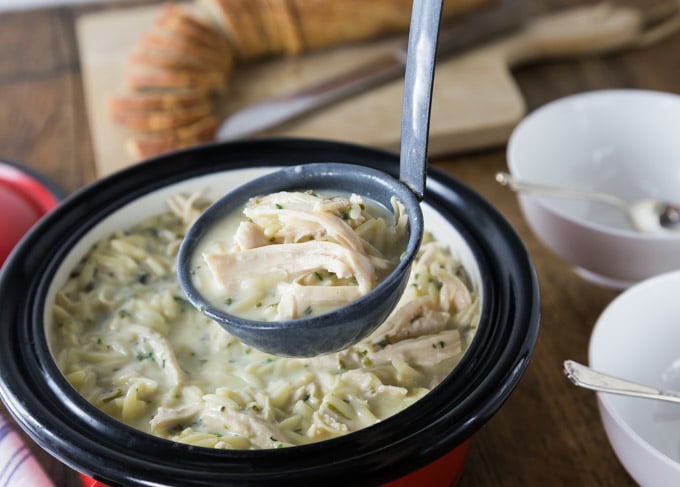 a ladle filled with chicken soup with bowls and spoons on the right and bread in the background