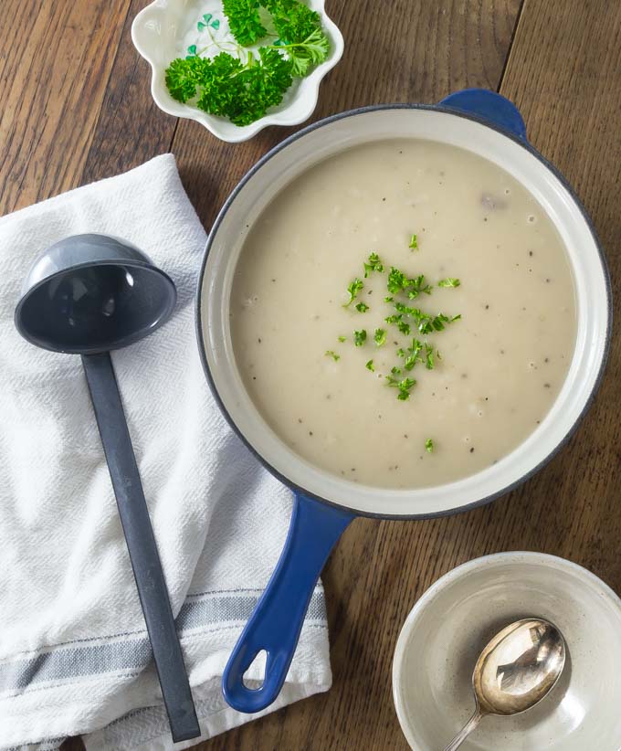 A blue dutch oven of Irish potato soup with parsley on top and a ladle on a napkin on the left