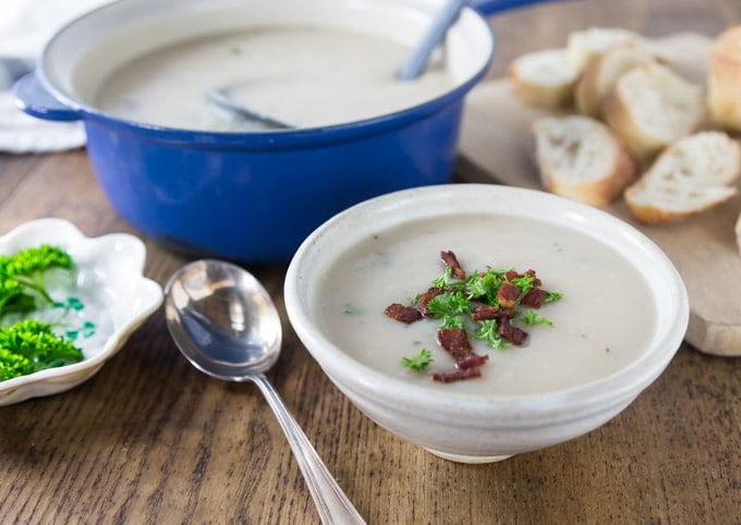 a bowl of potato soup topped with bacon and parsley with a spoon on the left and a pot of soup and bread in the back