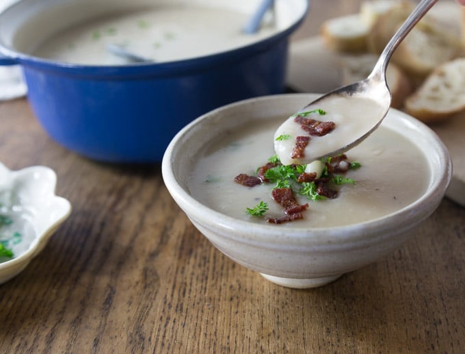 a spoon of potato soup with bacon and parsley over a bowl with a pot of soup and bread in the back