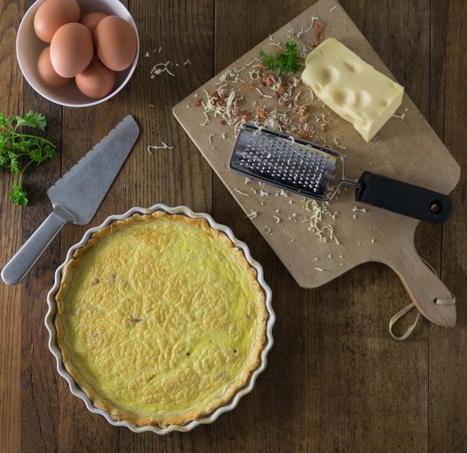 a quiche lorraine in a baking dish with a bowl of eggs, parsley, a cutting board with a block of cheese and a grater
