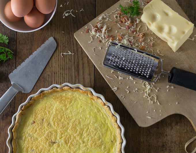 a close up of a quiche in a baking dish and a cutting board with grated cheese and a bowl of eggs