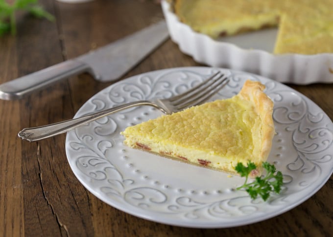 a slice of quiche on a white plate with a fork and a piece of parsley