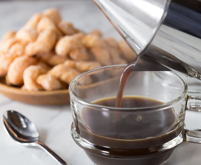 a glass cup of coffee being poured with snacks in the background