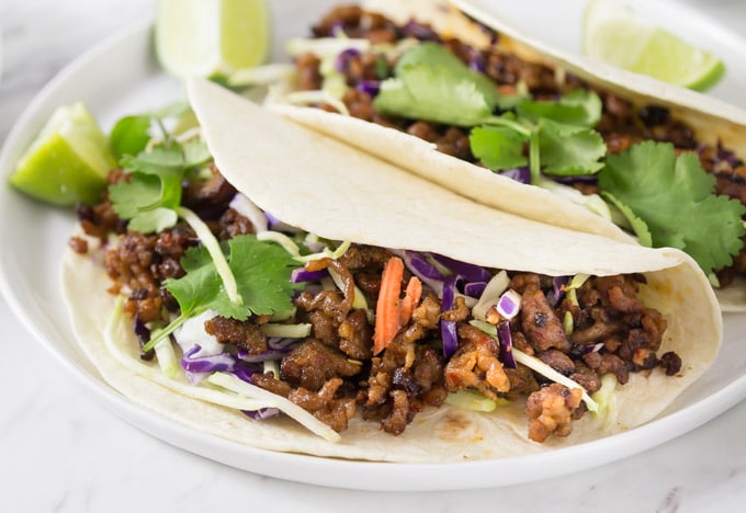 two tacos on a white plate with red cabbage, coriander, and lime wedges
