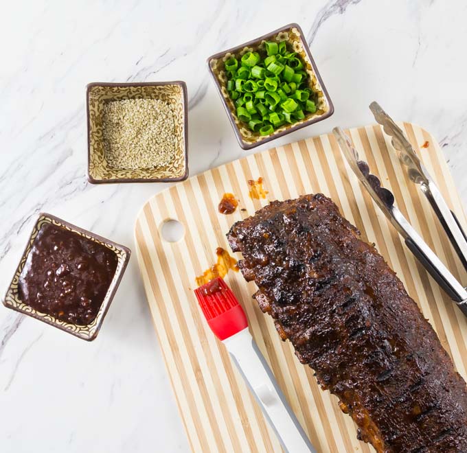 a cooked rack of ribs on a cutting board with bowls of spices, sauce, and green onion slices