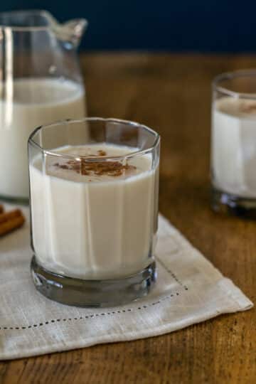 A photo of coquito in a cocktail glass with a glass pitcher in the background.
