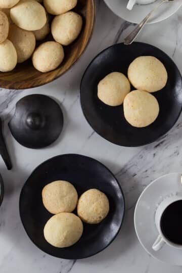 A photo of Brazilian cheese bread on a black plate with a wooden bowl and coffee cups.