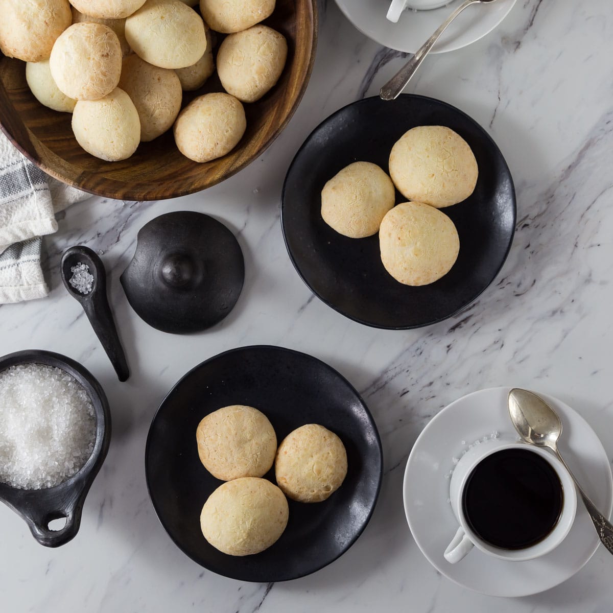A photo of Brazilian cheese bread on a black plate with a wooden bowl and coffee cups.