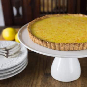 A lemon tart on a white serving plate with plates and forks in the background.