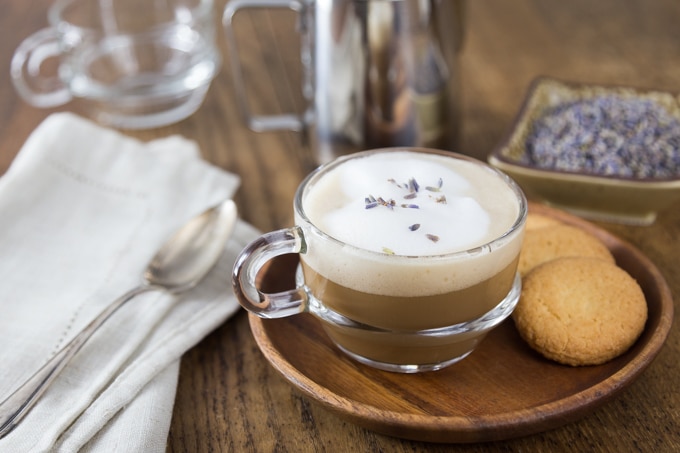 A photo of a clear coffee cup with lavender latte and some cookies.