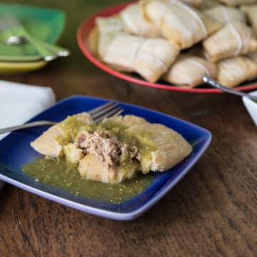 A photo of pork tamales on a blue plate with salsa verde.