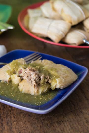 A photo of pork tamales on a blue plate with salsa verde.