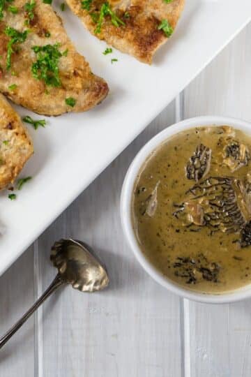 A photo of pork chops on a white plate next to a bowl with morel mushroom sauce.