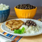 A photo of chuletas, black beans and rice on a white plate.
