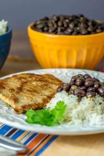 A photo of chuletas, black beans and rice on a white plate.