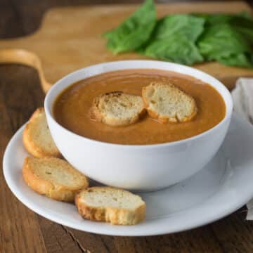 A white bowl with tomato basil bisque with toasted baguette slices.