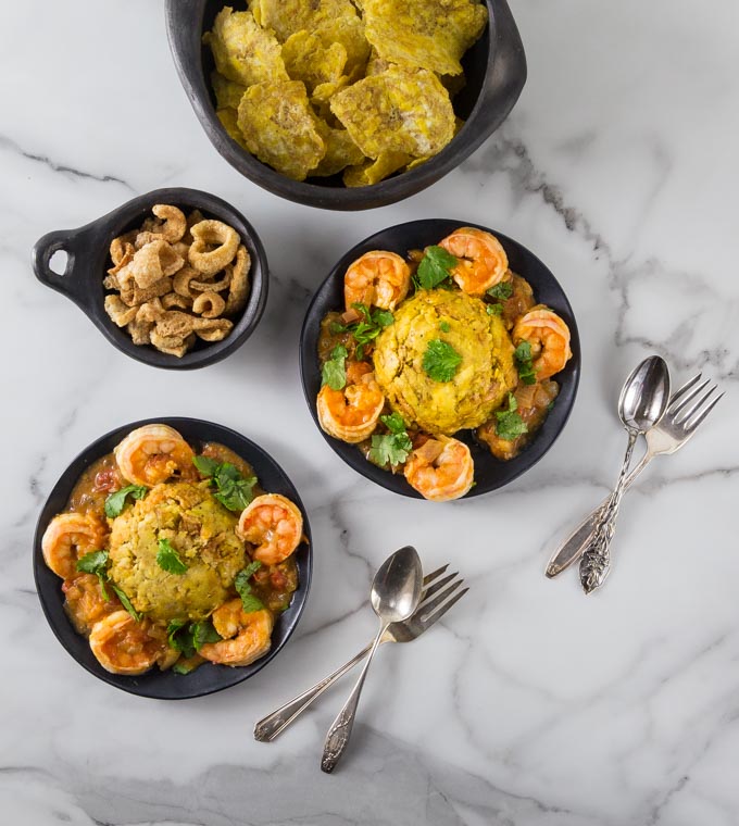 An overhead photo of plates with shrimp, mofongo along with tostones and plantain chips.