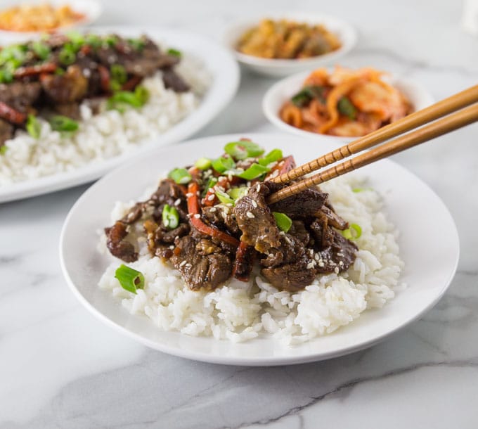 A photo of a white plate with rice and beef bulgogi and chop sticks picking up a piece of beef.