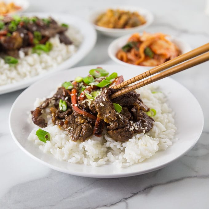 A white plate with rice and Korean bulgogi beef being picked up with a chop stick.