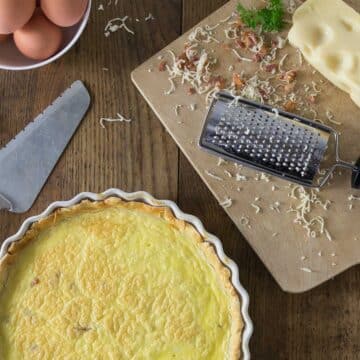 A photo of quiche lorraine in a baking dish with a cutting board and grated cheese.