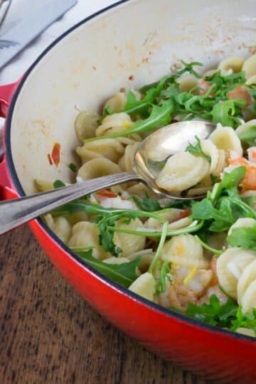 A photo of orecchiette paste with shrimp, fennel and arugula in a red pan with a spoon.
